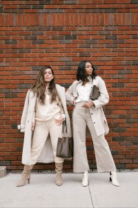 Two women in cream outfits against brick wall.